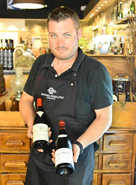 Yann au bistrot, en tablier, présentant deux bouteilles